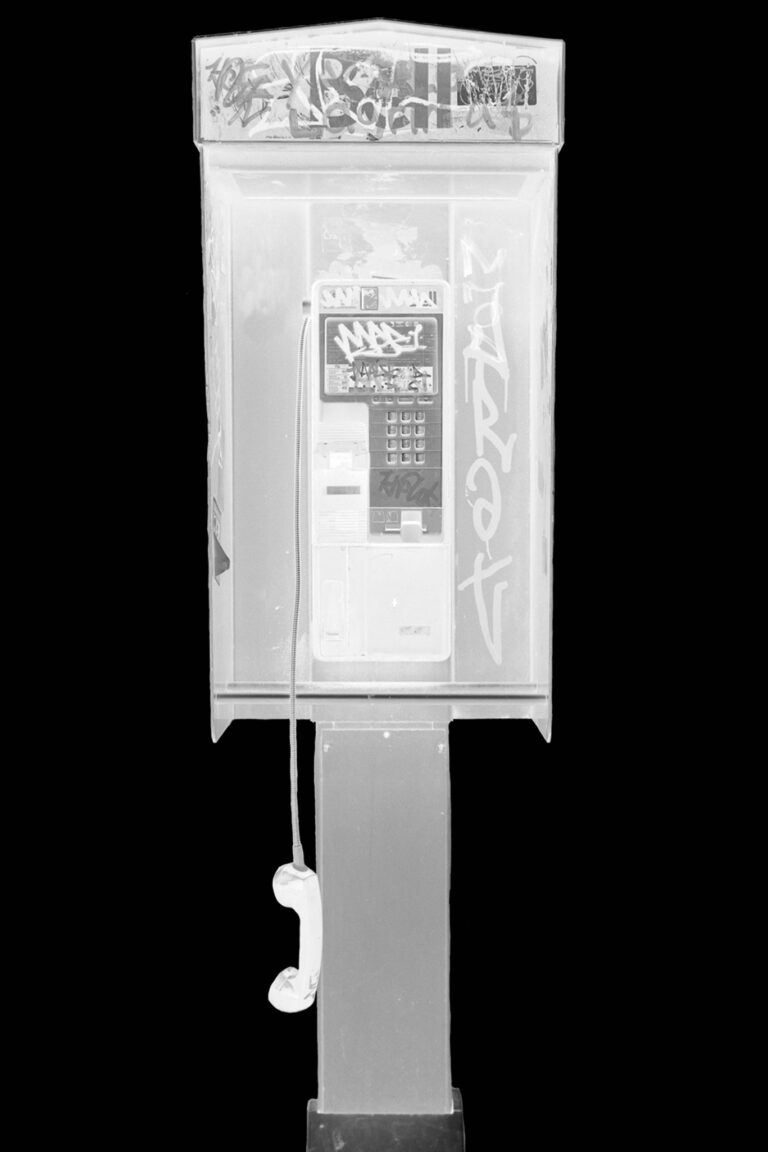 A black and white photo of a pay phone with a thick black border around it. The payphone has graffiti on the top and sides around the phone. The phone is hanging down off its stand by the cord.