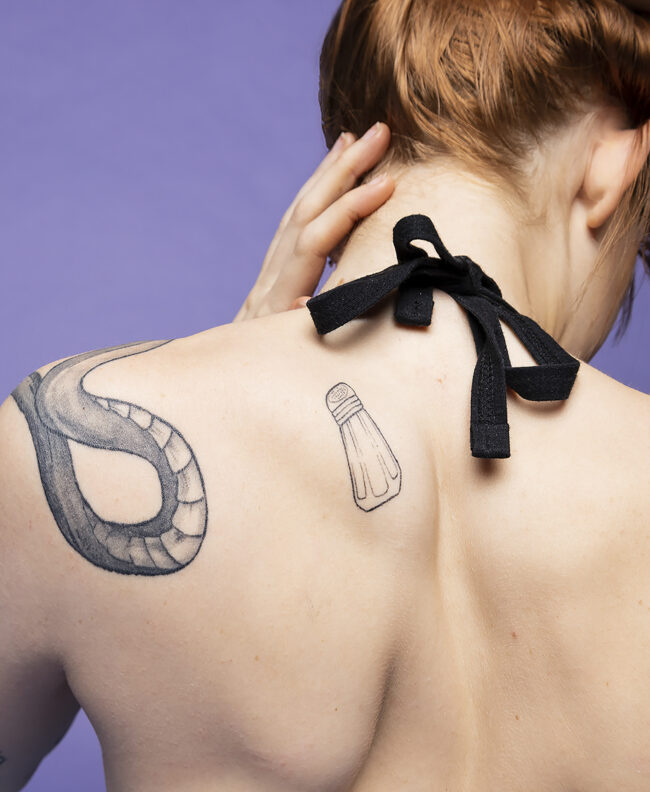 A photo of a womens back, featuring two tattoos of a salt shaker and snake, the woman's hand caresses the back of her neck