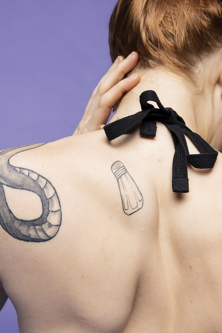 A photo of a womens back, featuring two tattoos of a salt shaker and snake, the woman's hand caresses the back of her neck