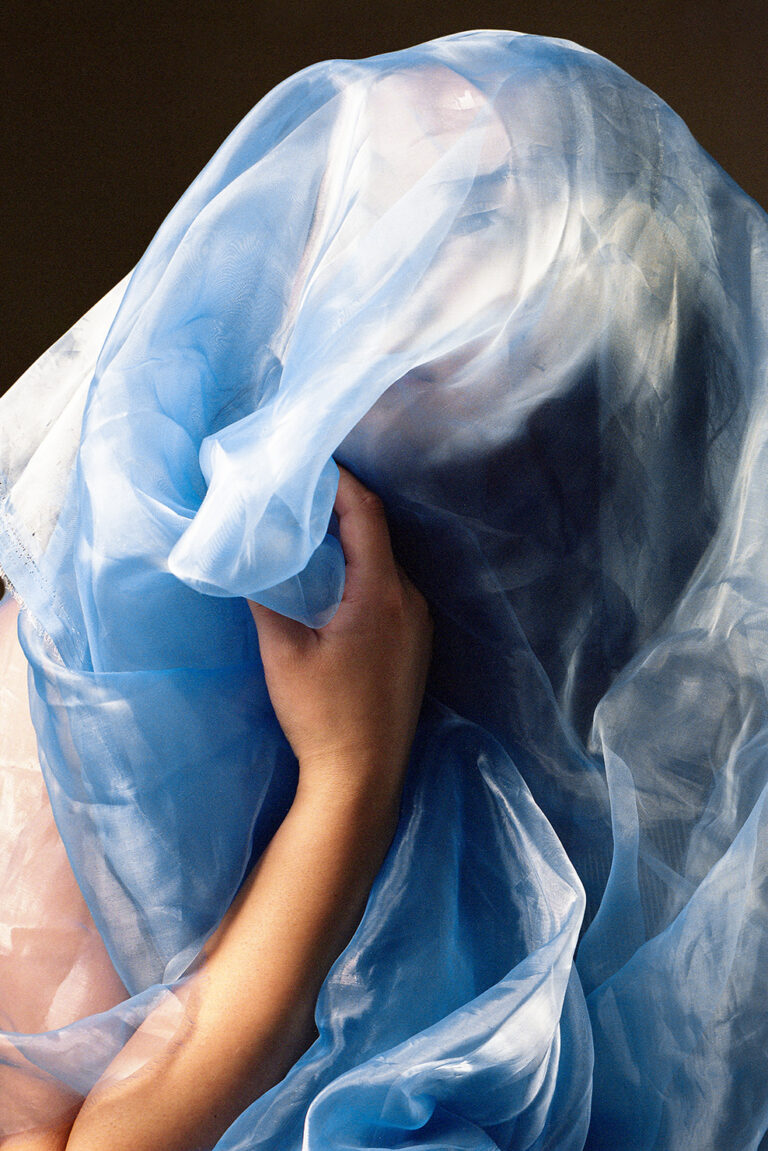 A photograph of someone wrapped in a blue sheer fabric, their hand grasps the fabric and their face is partially visible behind the fabric