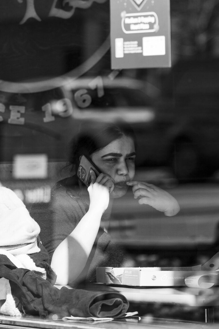 black and white image of a woman on a call while grabbing food shot through a window
