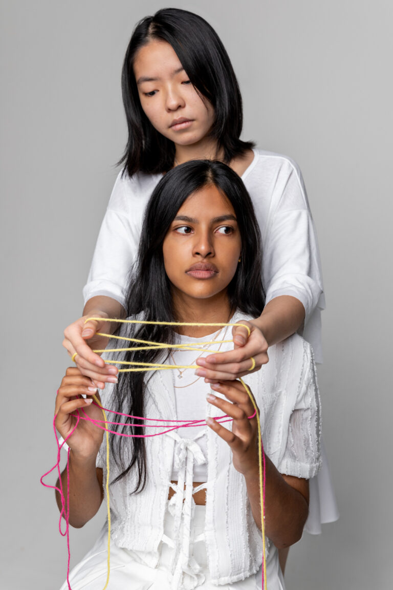 Two women, one sitting the other standing with her arms over the shoulders of the other. Their hands are intertwined with yarn looking away and at their hands. Both women are wearing white and the yarn is both yellow and pink.