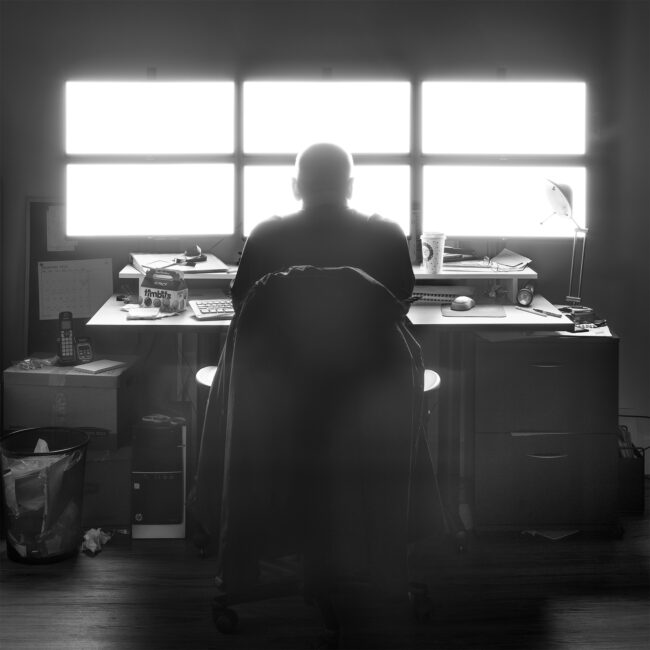 A security attendant sits at a desk awash in the light from an array of six monitors.