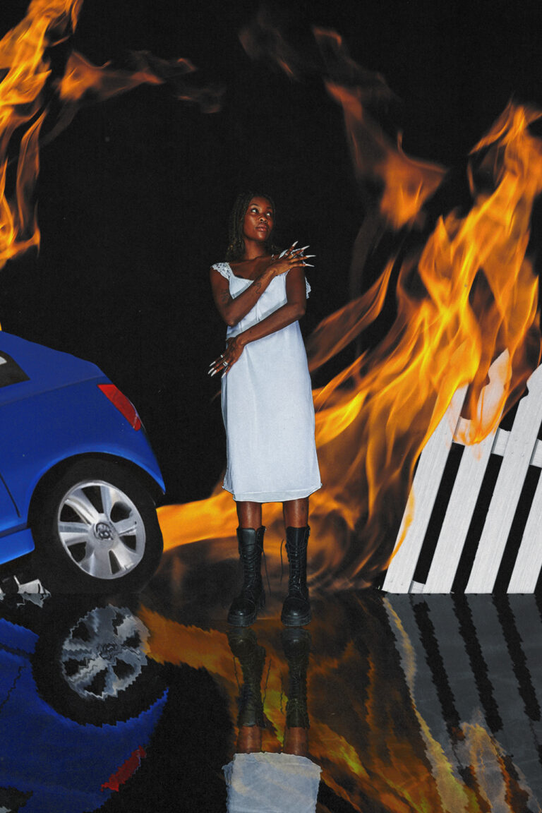 A person stands in the middle of the photograph while being surrounded by flames, to the left of them is the back end of a car and to the right of them a white picket fence. The bottom quarter of the image is reflected