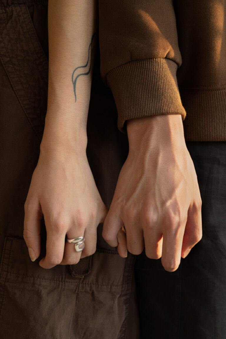 This photograph is a close up detail shot of two people's hands linked by their pinkies. The hand to the left is wearing a silver ring on their middle finger. The hands are at waist level and the overall colour scheme consists of warm browns and greys.