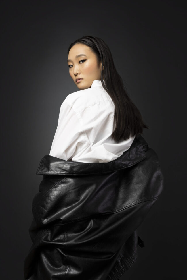 Asian model standing looking over her shoulder wearing a white shirt and black leather jacket against a dark backdrop.