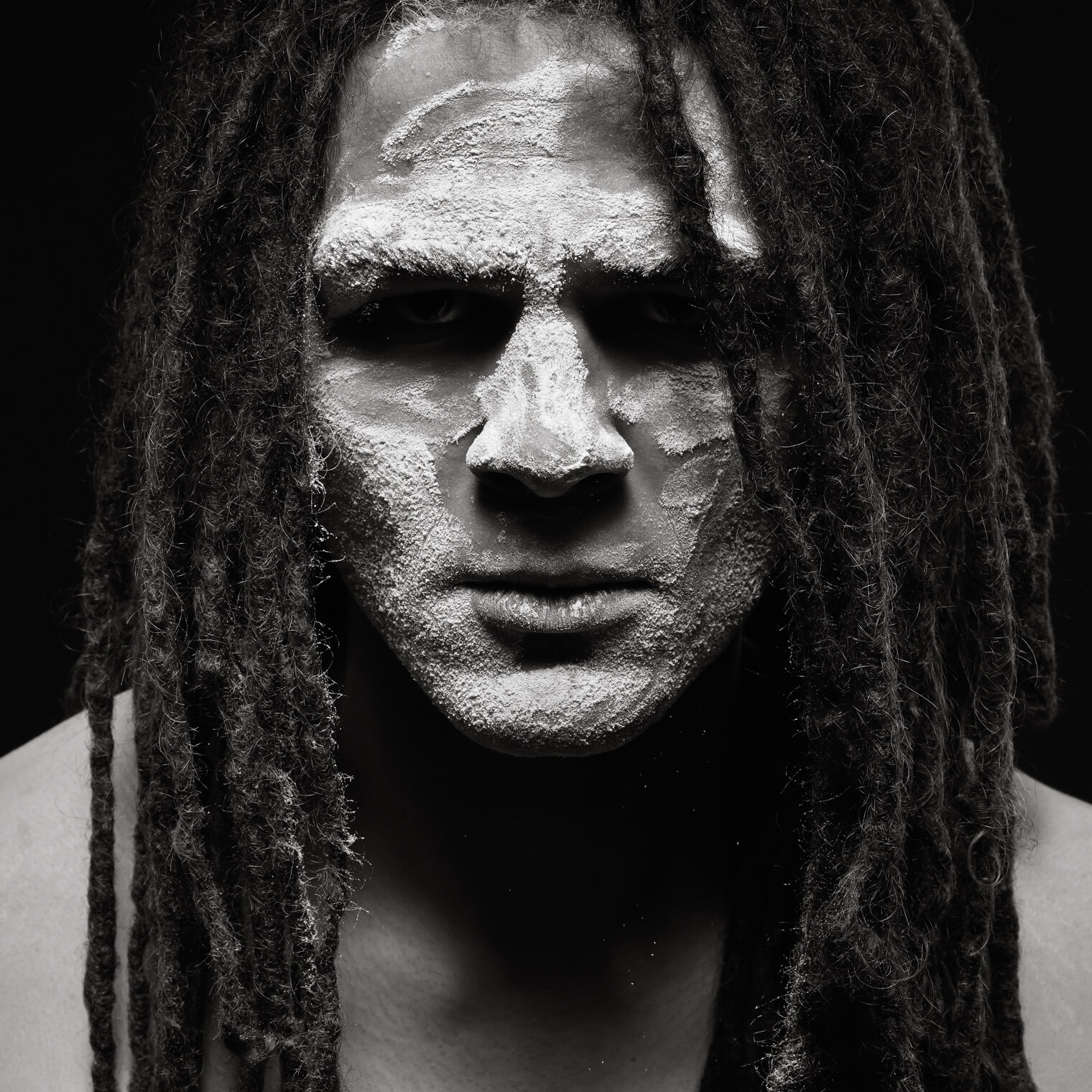 A man with long hair in dreadlocks is facing the camera head on, gazing into the lens. His face is covered with a white powder as some falls off his face the moment the photo was taken.