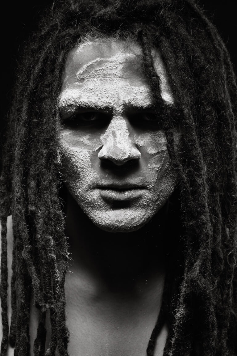 A man with long hair in dreadlocks is facing the camera head on, gazing into the lens. His face is covered with a white powder as some falls off his face the moment the photo was taken.