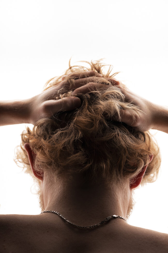 The back of someone's head leaning partially forward with their hands in their blond hair. The subject is back lit and a chain sits upon their neck.