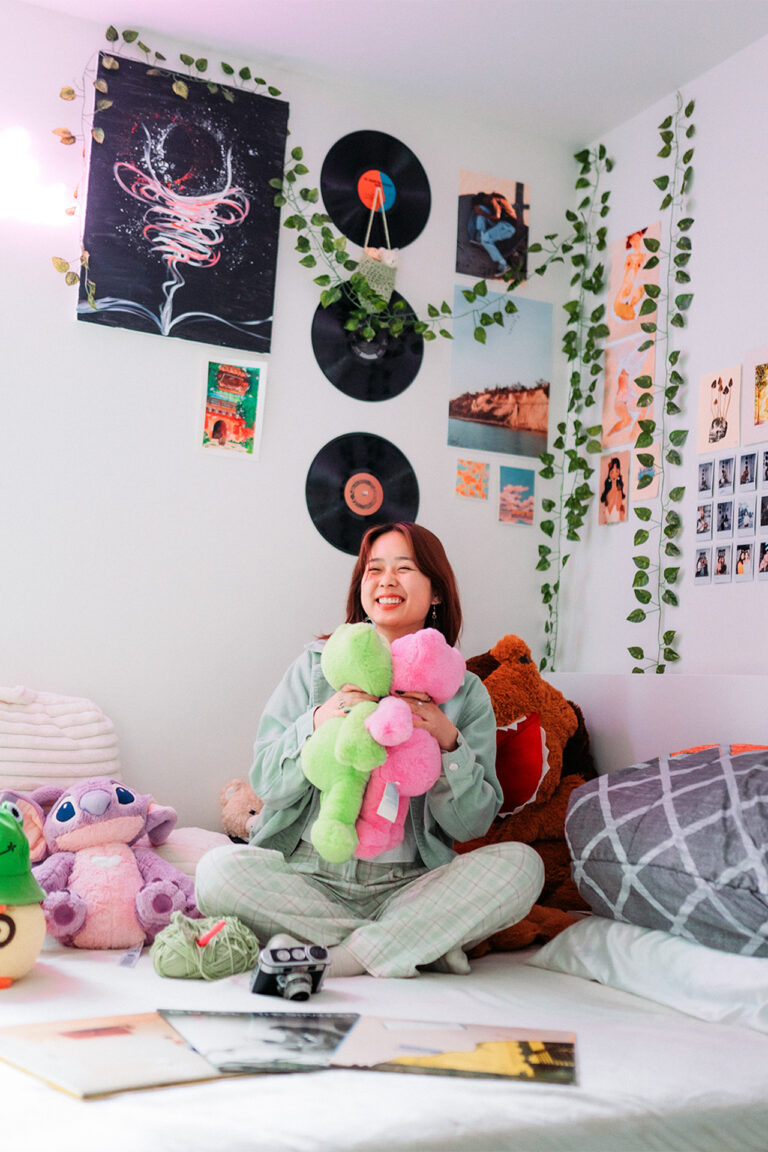 A portrait of a someone surrounded by stuffed animals and various objects of their room, they sit on their bed and smile while holding two stuffed animals in both hands