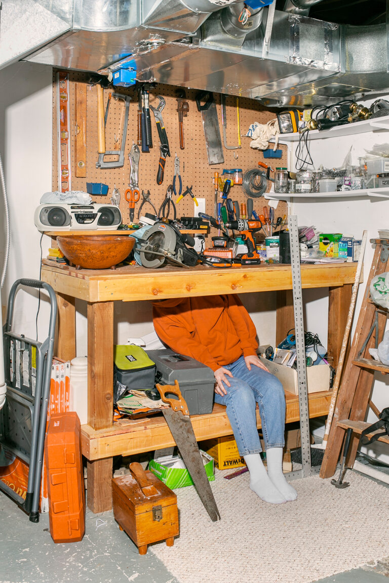 A person sits on a shelf of a wooden workbench, awkwardly hiding their face from view. There are a various tools hanging on the wall about the bench, as well as spread out on the surface of the bench. There is a saw leaning against the shelf on the floor. A metallic vent runs through the top of the frame.