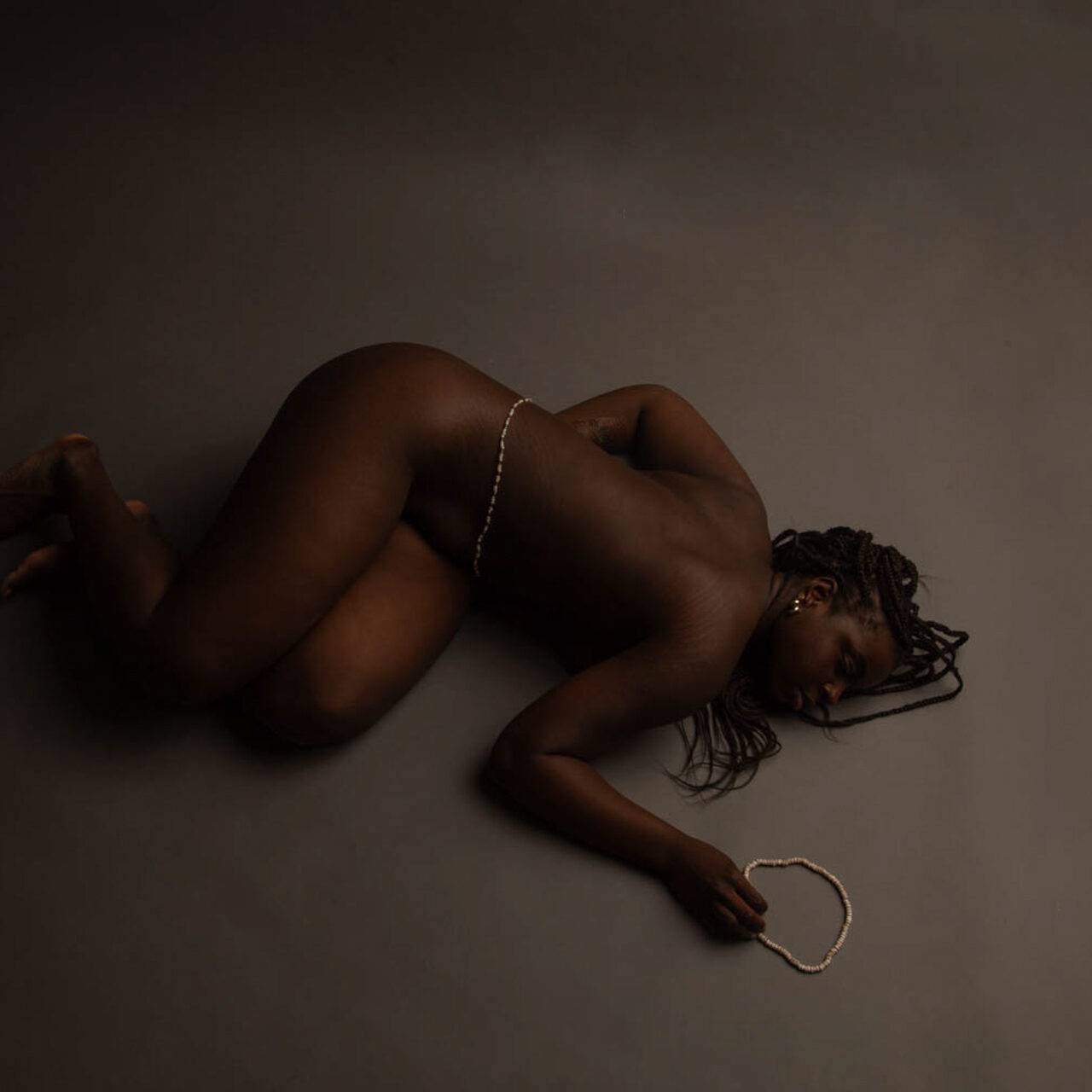 The photo consists of a Black woman laying on the ground nude with white African waist beads on her waist and laying next to her hand.