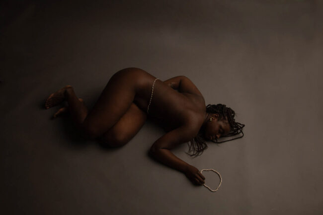 The photo consists of a Black woman laying on the ground nude with white African waist beads on her waist and laying next to her hand.