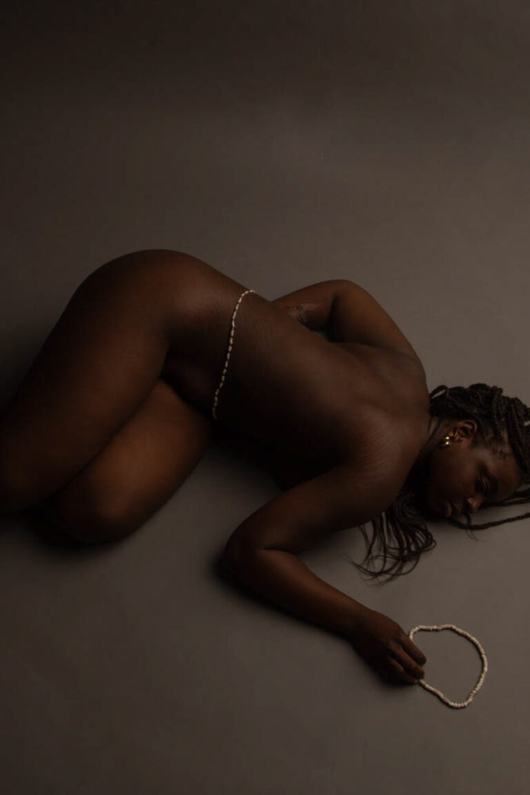 The photo consists of a Black woman laying on the ground nude with white African waist beads on her waist and laying next to her hand.