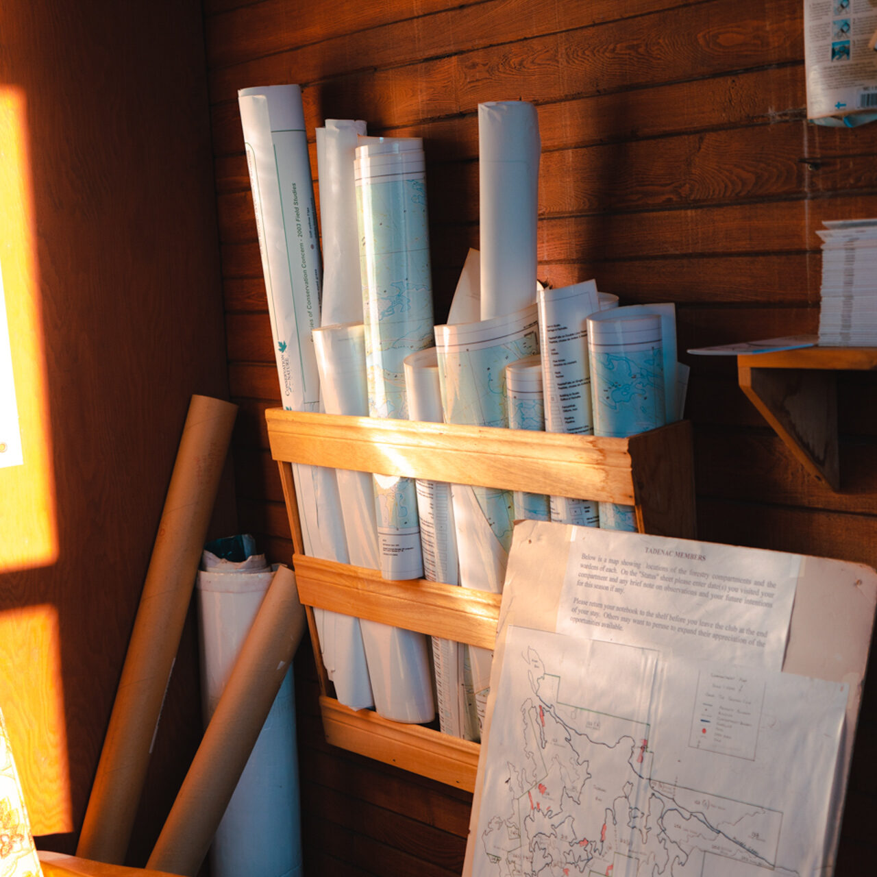 This photo consists of a brightly lit wooden cabin room. There are several maps rolled up in the corner of the room and stored on a hanging shelf. There is also a map on a wooden stand with colored outlines and descriptions.