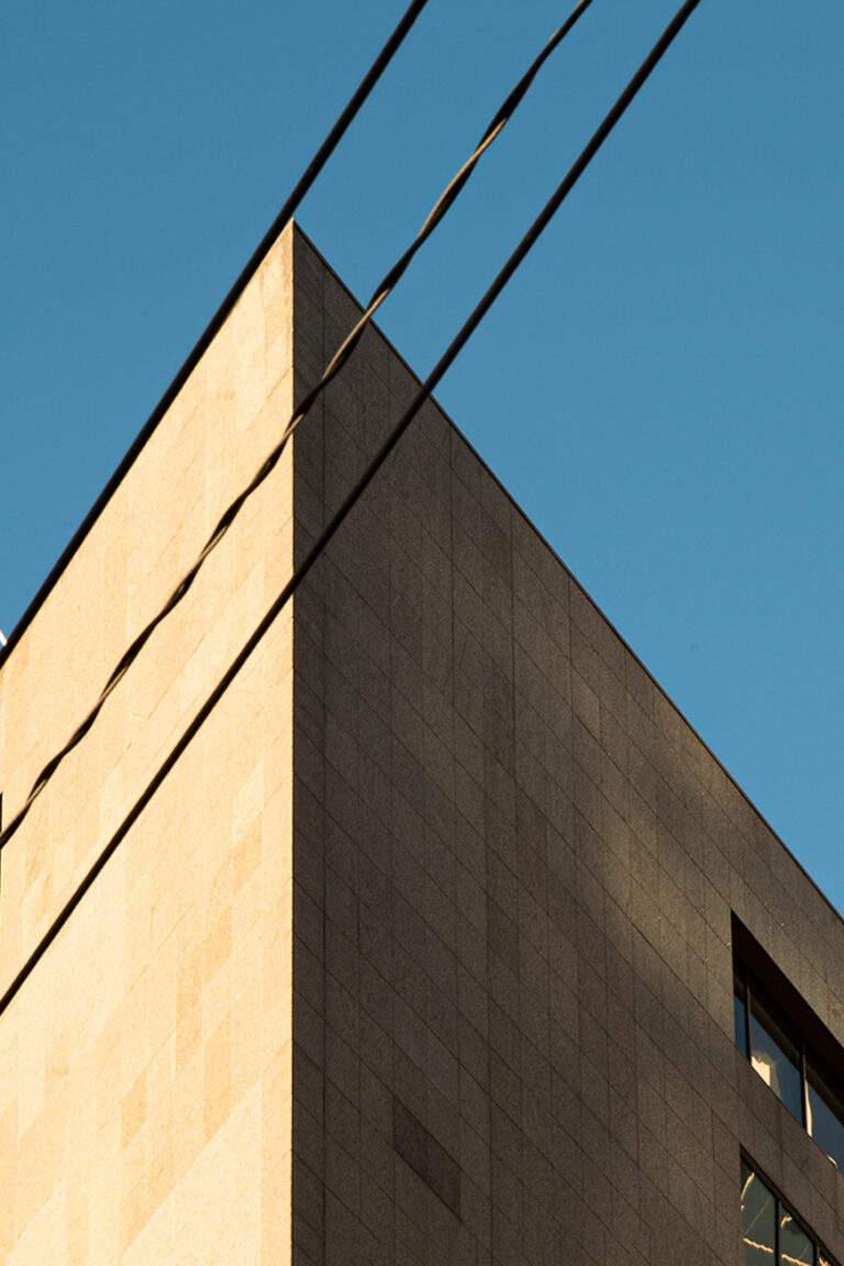 A photo looking up at a corner of a building, three power lines align with the corner of the building diagonally across the image. The sky is a bright blue that matches well with the beige exterior of the building.