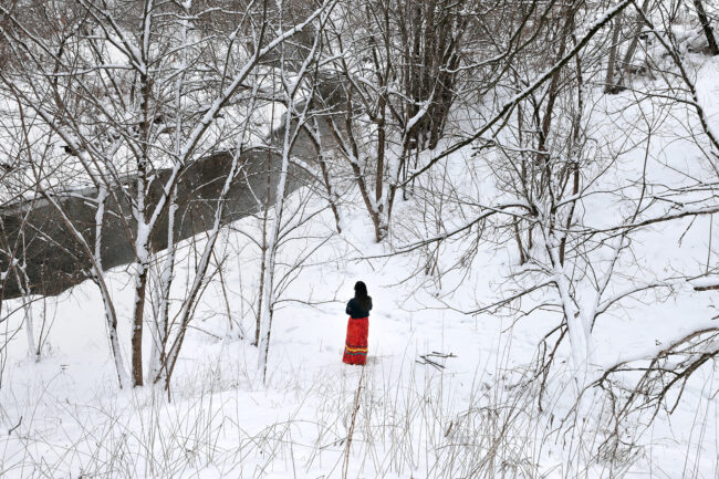 A landscape image of a forest and a creek covered in snow taken. The image has been taken from uphill, giving an expansive look into the forest. The subject is in the center of the image, closer to the bottom. They are surrounded by trees and branches but are visible in a small clearing. Their back is facing the camera and they are wearing a black long-sleeve shirt and an orange patterned ribbon skirt with three green and yellow ribbons wrapping around the bottom half of the skirt. The creek is visible from the bottom left corner of the image and trails up towards the center.