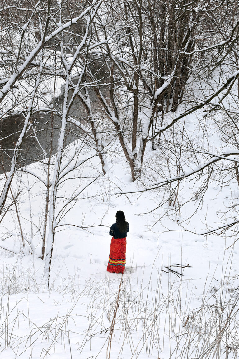A landscape image of a forest and a creek covered in snow taken. The image has been taken from uphill, giving an expansive look into the forest. The subject is in the center of the image, closer to the bottom. They are surrounded by trees and branches but are visible in a small clearing. Their back is facing the camera and they are wearing a black long-sleeve shirt and an orange patterned ribbon skirt with three green and yellow ribbons wrapping around the bottom half of the skirt. The creek is visible from the bottom left corner of the image and trails up towards the center.