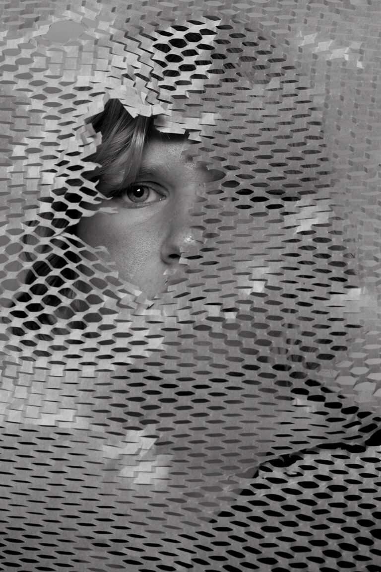 This is a black and white photo of a girl at a close up angle. She is standing behind a crinkled sheet of honeycomb packing paper which obscures most of her face. There is a hole torn out of the paper revealing one of the girl's eyes with a portion of her cheek and nose.
