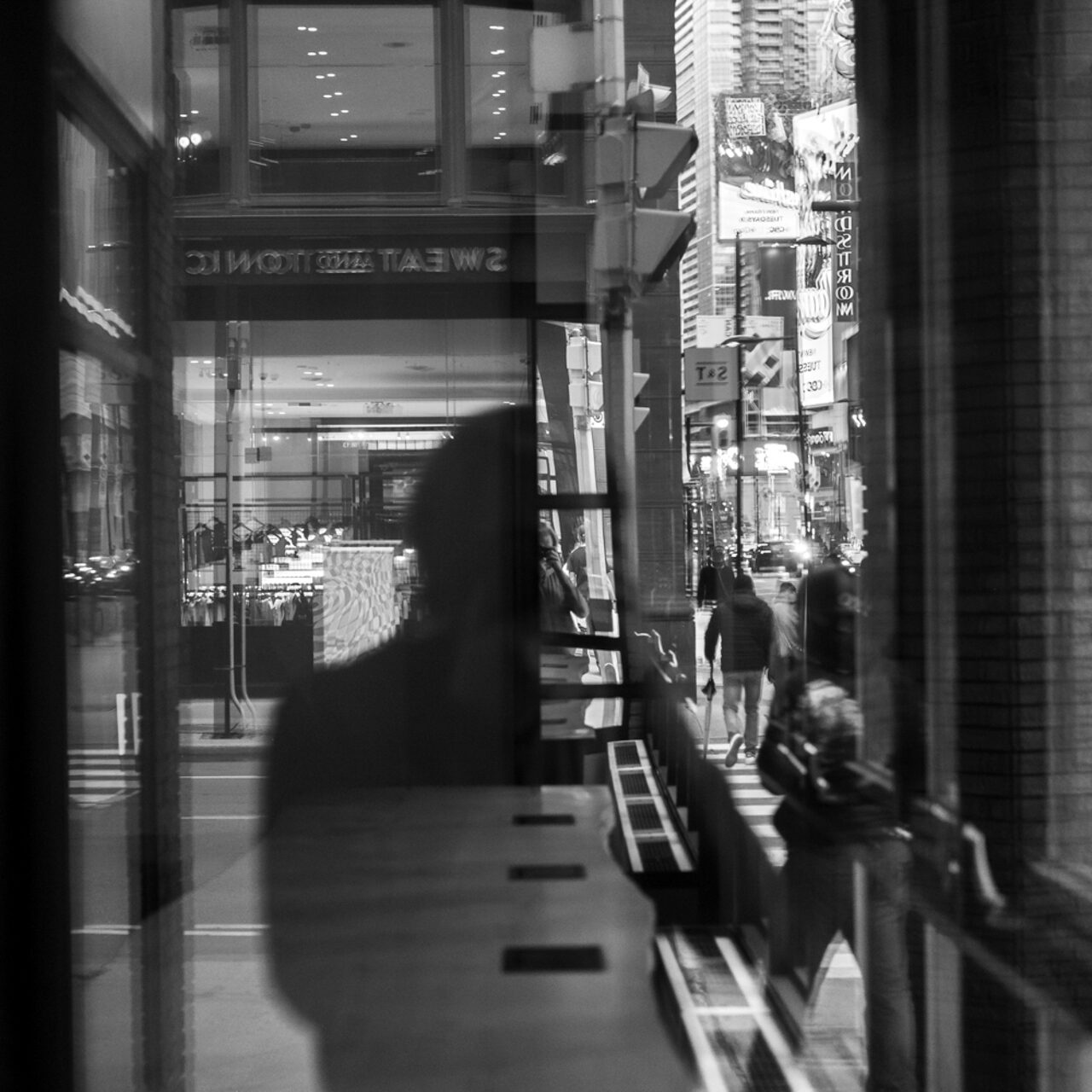 This photo consists of the reflection of a man’s silhouette and the streetscape with several moving people and cars on the right side of the photo.