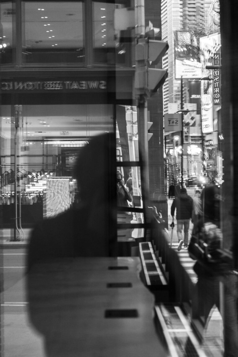 This photo consists of the reflection of a man’s silhouette and the streetscape with several moving people and cars on the right side of the photo.