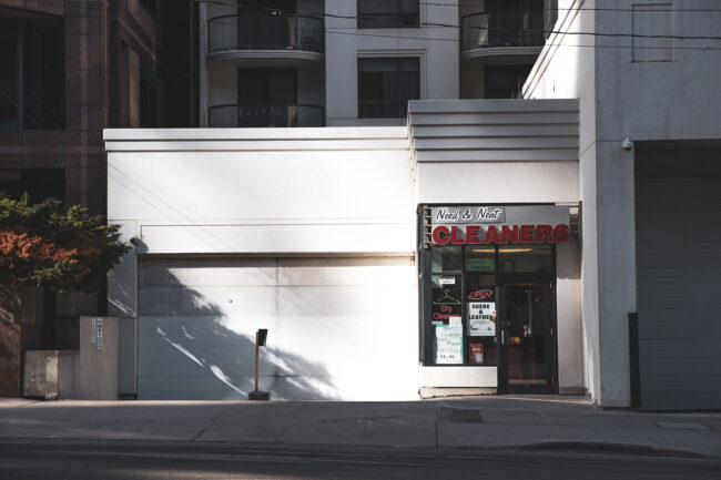 A small, white building with a red "Cleaners" sign. The building is partially cast in shadow from a tree pictured on the left side of the frame. There are apartment building windows seen above as well as to the left of the cleaners.