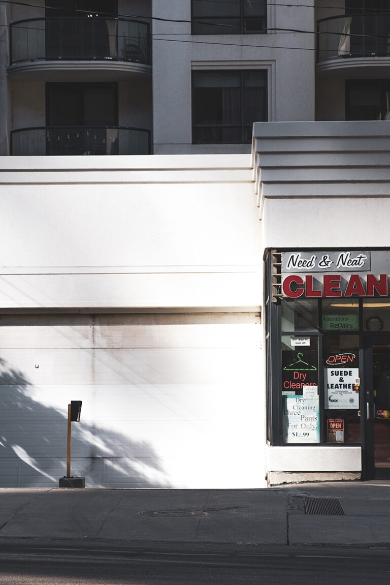 A small, white building with a red "Cleaners" sign. The building is partially cast in shadow from a tree pictured on the left side of the frame. There are apartment building windows seen above as well as to the left of the cleaners.