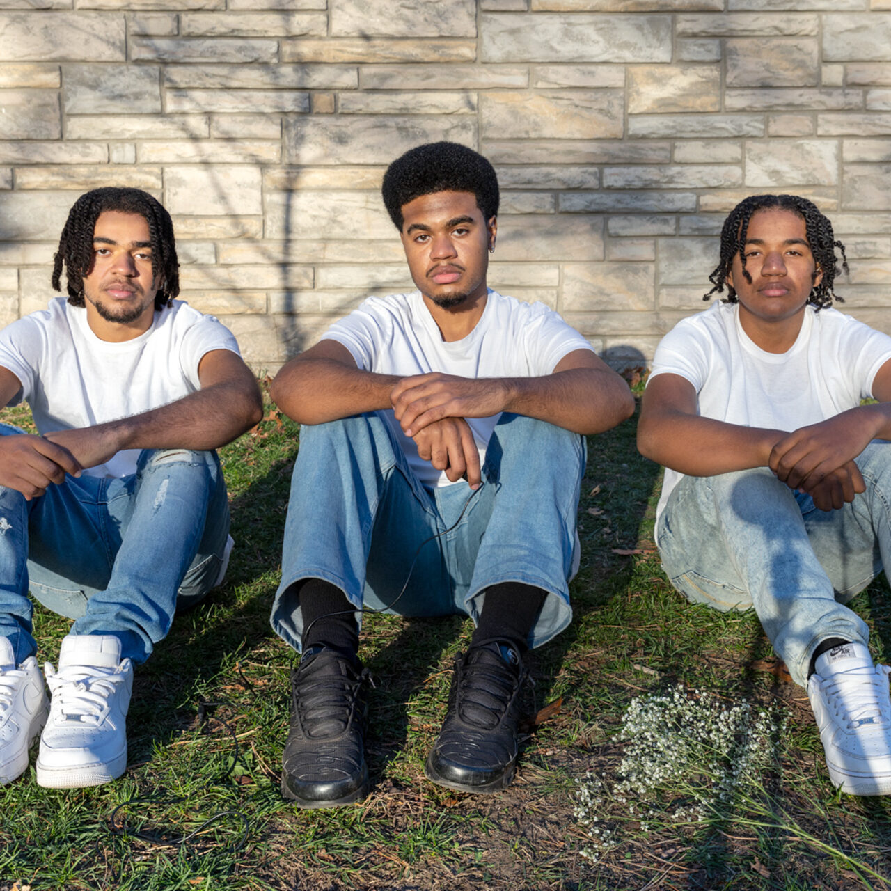 This photo consists of three Black men who are sitting in a grass field on a bright day, all gazing into the camera. They are all wearing white short-sleeved shirts and light-blue jeans. They wear alternating color shoes; white, black, white. The same goes for their hair, the first is twisted, the second is an afro and the last is twisted as well. The three of their arms are crossed over their knees, the man in the middle holds a black shutter release cable and a bunch of Baby’s Breath flowers lay to the right of him.