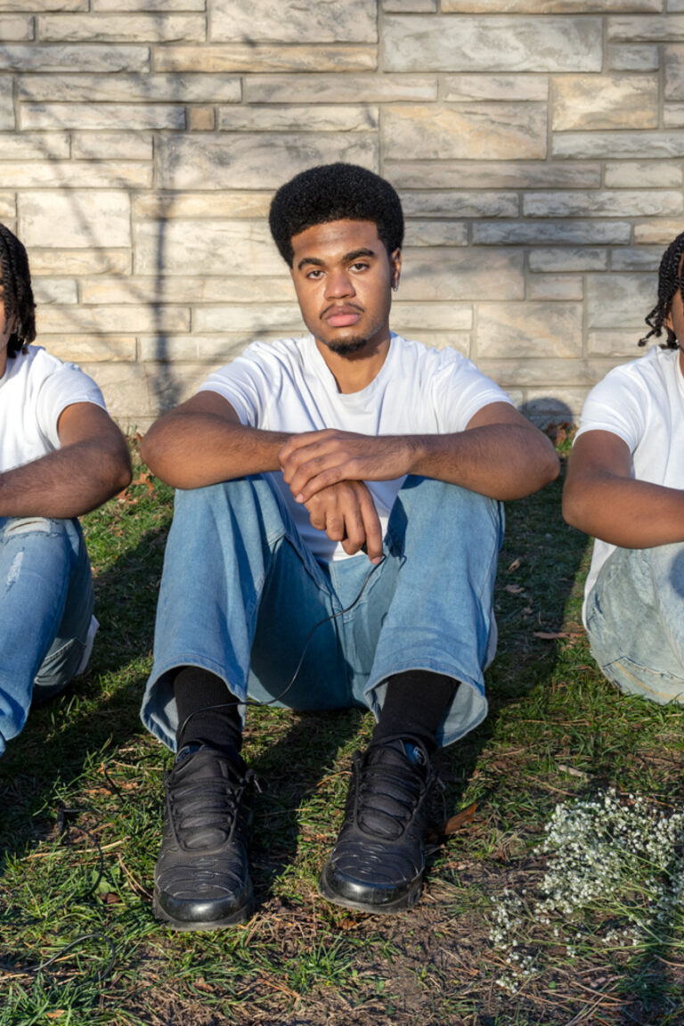 This photo consists of three Black men who are sitting in a grass field on a bright day, all gazing into the camera. They are all wearing white short-sleeved shirts and light-blue jeans. They wear alternating color shoes; white, black, white. The same goes for their hair, the first is twisted, the second is an afro and the last is twisted as well. The three of their arms are crossed over their knees, the man in the middle holds a black shutter release cable and a bunch of Baby’s Breath flowers lay to the right of him.