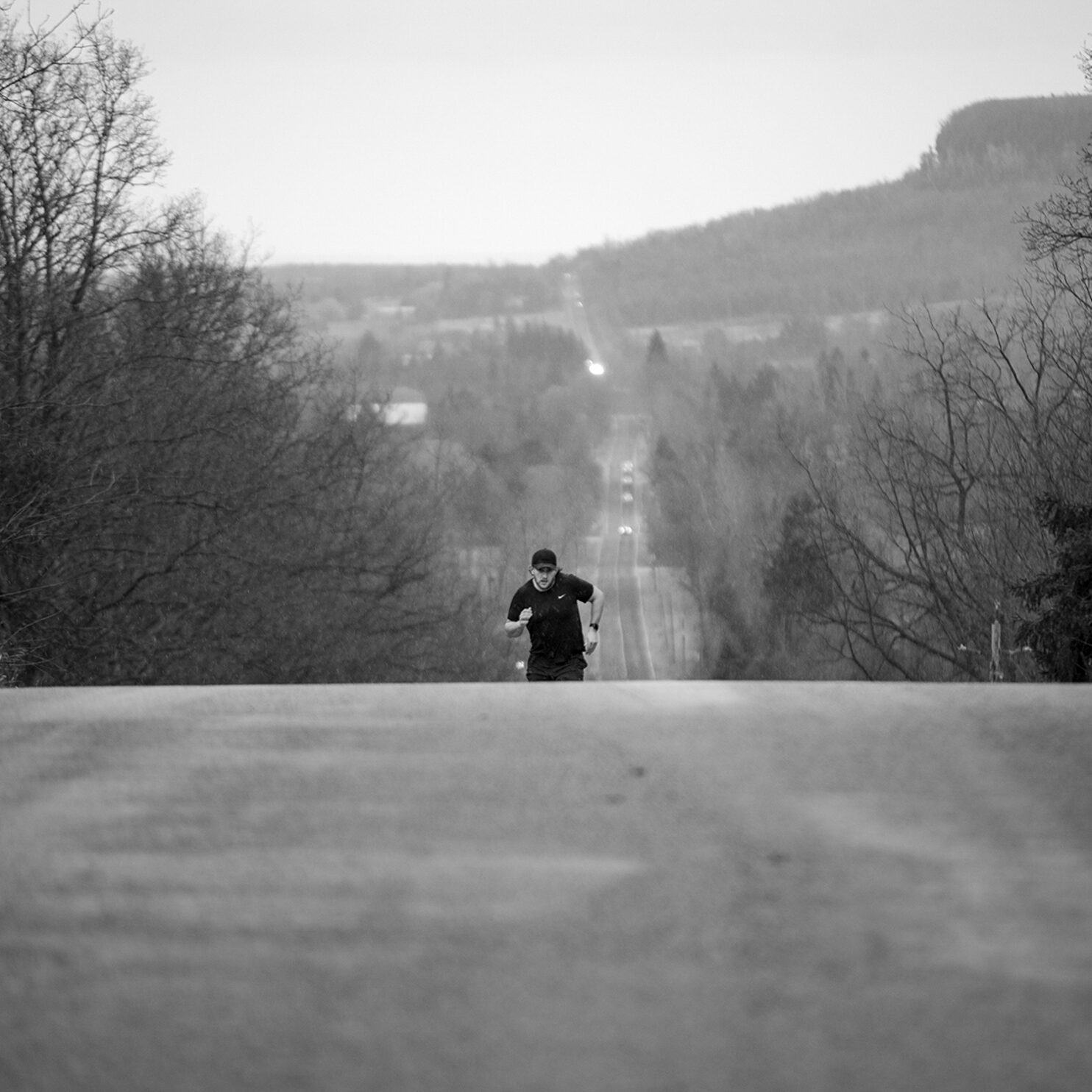 The photo depicted is in black and white. A man is running up a steep road. He is wearing all black and is captured striding up the road free of cars.