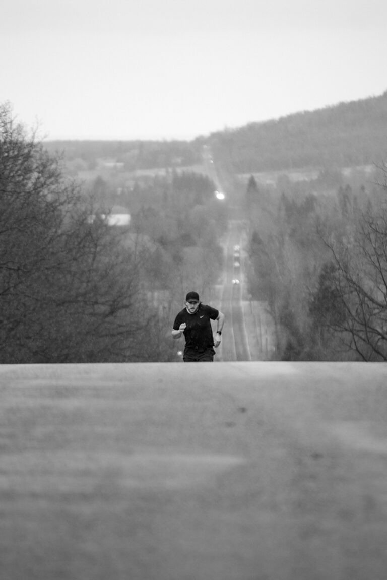The photo depicted is in black and white. A man is running up a steep road. He is wearing all black and is captured striding up the road free of cars.
