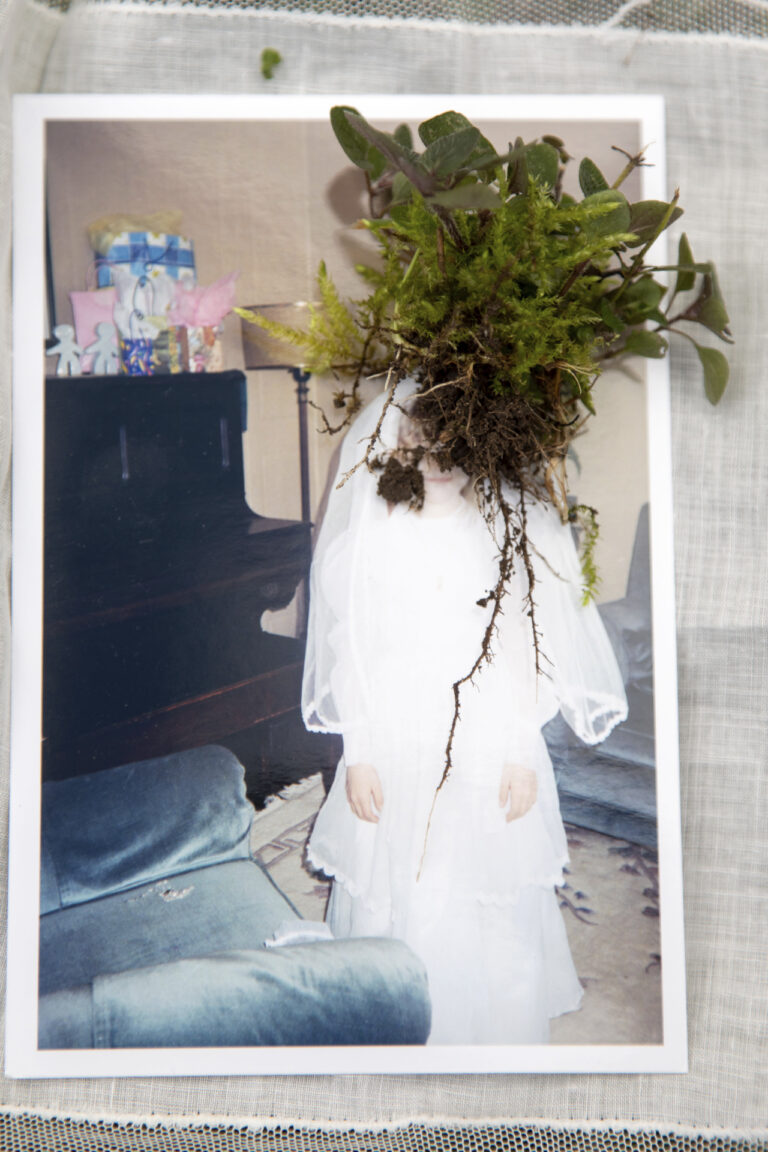 an image of a young girl in a communion dress sitting on top of a white lace doily. There is an uprooted weed placed on top of the image over the girl's face.