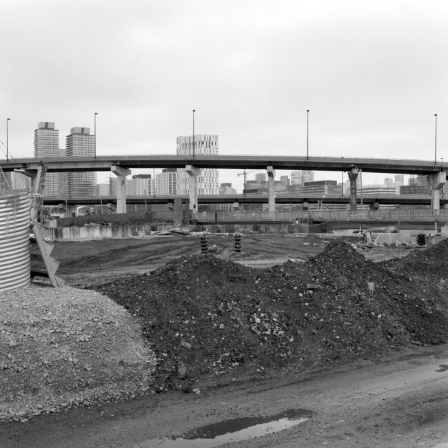 Where The River Runs # 1: A square black and white photograph taken from a construction site. An embankment of dirt and gravel cuts through the middle of the frame. A highway can be seen beyond, with one pillar reading Don River. High-rise buildings also sprout up in the distance.