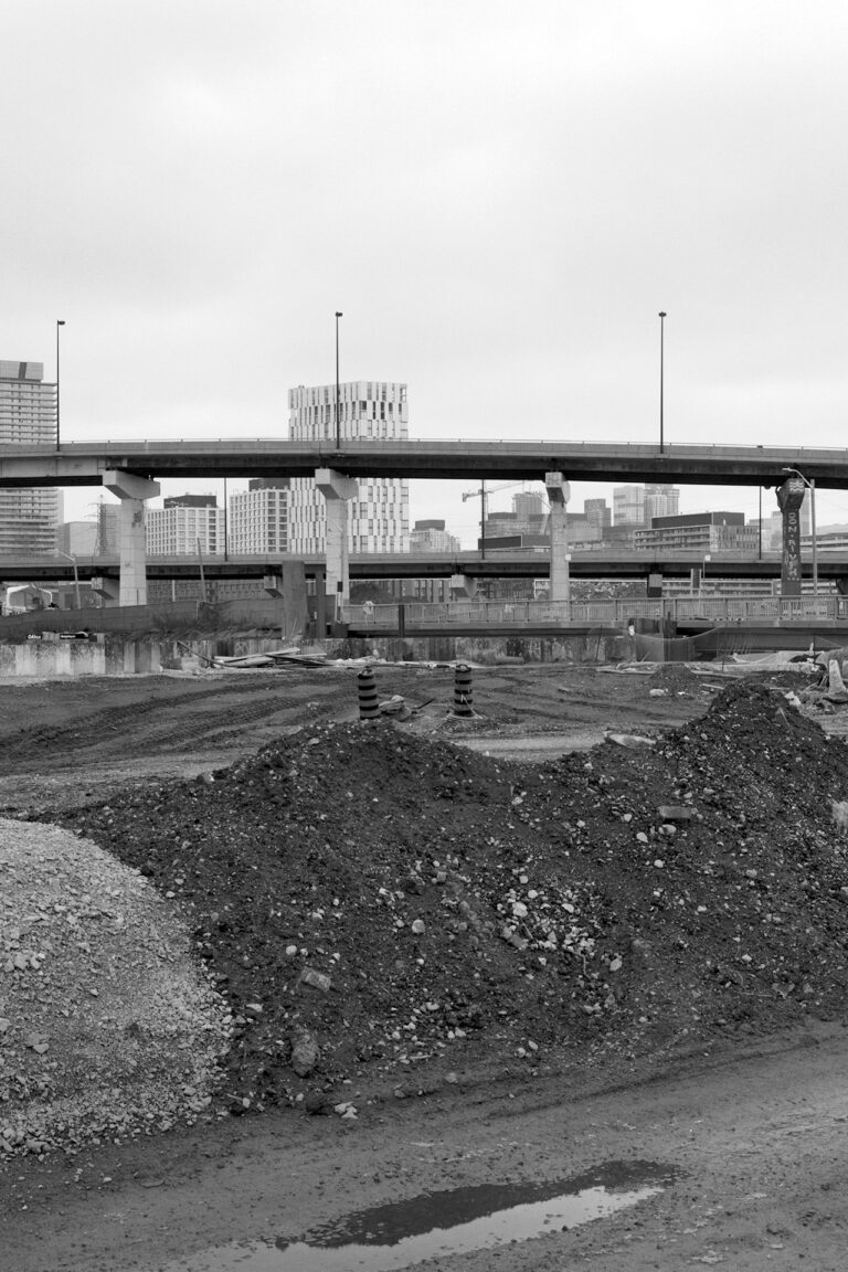 Where The River Runs # 1: A square black and white photograph taken from a construction site. An embankment of dirt and gravel cuts through the middle of the frame. A highway can be seen beyond, with one pillar reading Don River. High-rise buildings also sprout up in the distance.