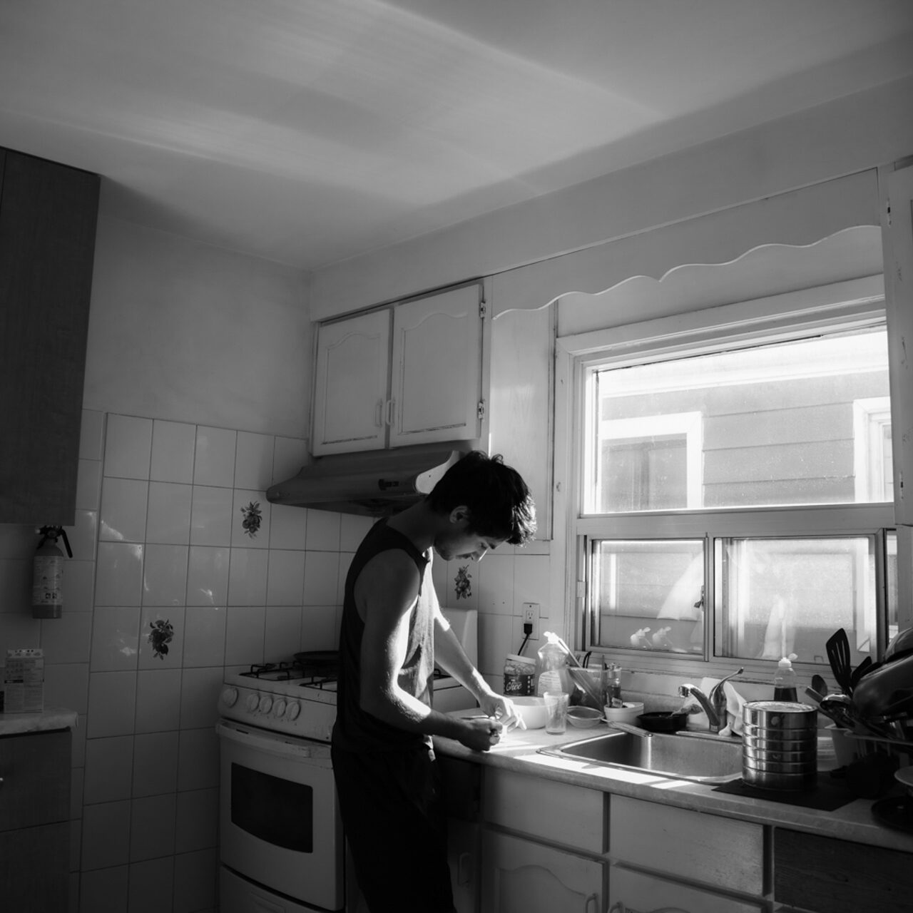 This photo consists of a man in a brightly lit kitchen preparing something to eat. He has short black hair and is wearing a sleeveless tank-top and pants. The photo is black and white.