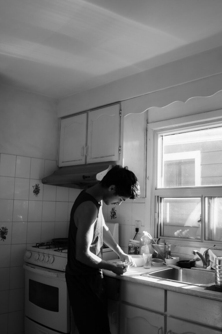 This photo consists of a man in a brightly lit kitchen preparing something to eat. He has short black hair and is wearing a sleeveless tank-top and pants. The photo is black and white.