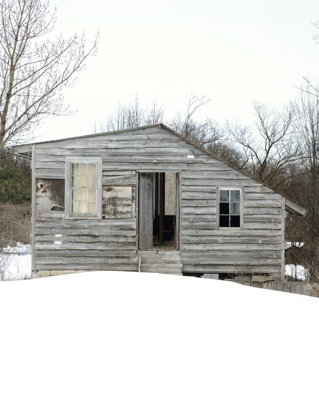 Untitled #3: An abandoned wooden cabin with broken windows and a slightly open door. It sits on a snowy plot of land with trees behind it.