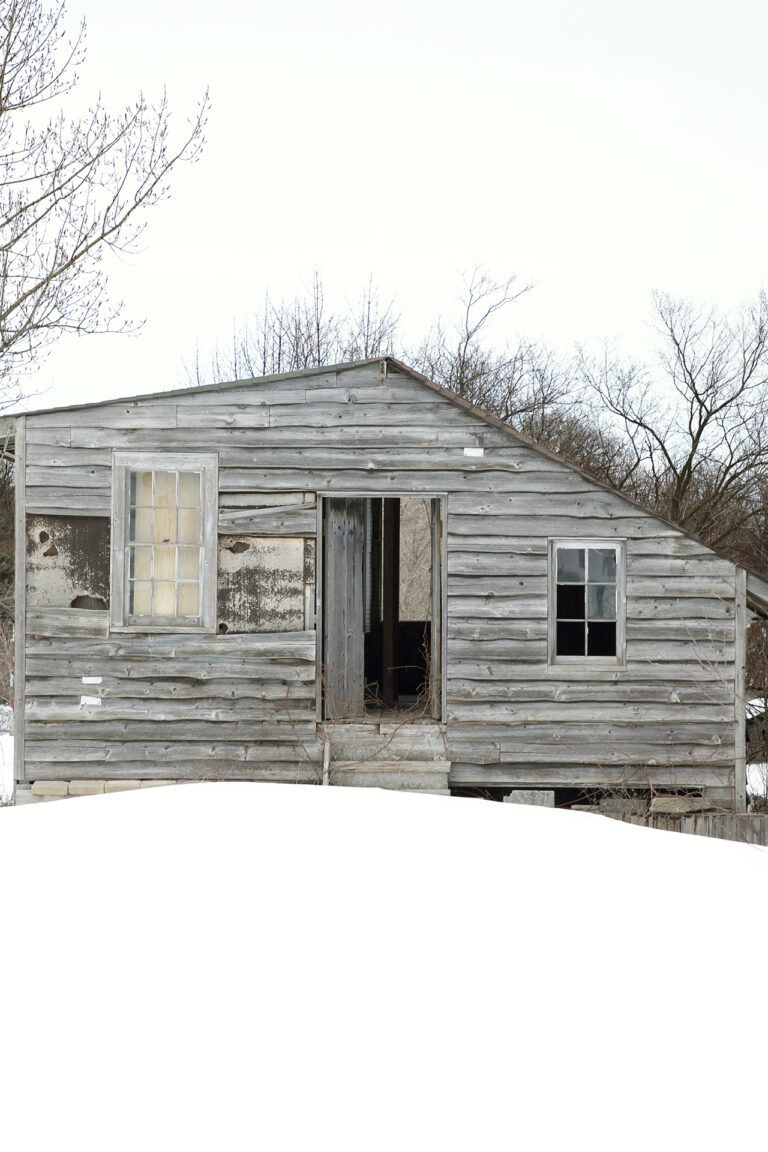 Untitled #3: An abandoned wooden cabin with broken windows and a slightly open door. It sits on a snowy plot of land with trees behind it.