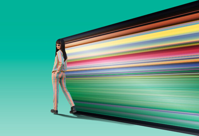 Model in a rainbow suit and dress pants walking away from the frame, the background a light deep turquoise green. As the model walks away from the edge, the colours from the subject are dragged out in a straight direction, exposing the incredible rainbow colours of the clothes. The clothing was supplied by (KnockTwiceToronto clothing)