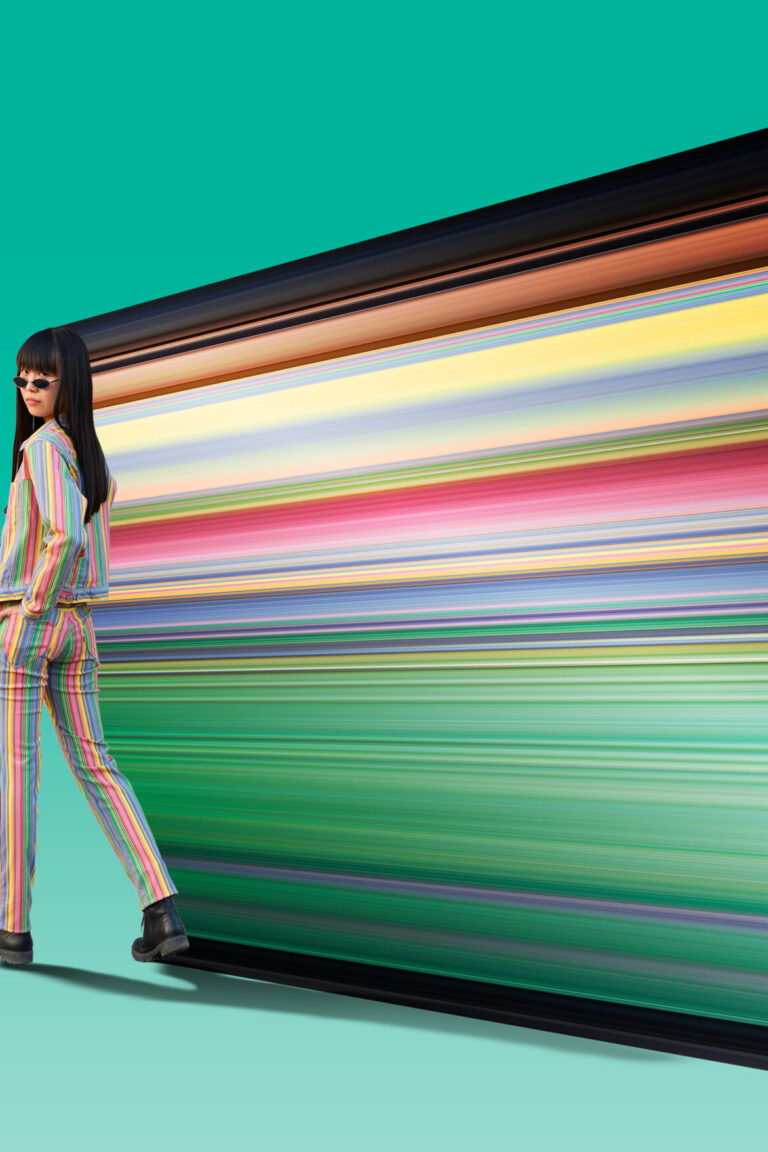 Model in a rainbow suit and dress pants walking away from the frame, the background a light deep turquoise green. As the model walks away from the edge, the colours from the subject are dragged out in a straight direction, exposing the incredible rainbow colours of the clothes. The clothing was supplied by (KnockTwiceToronto clothing)