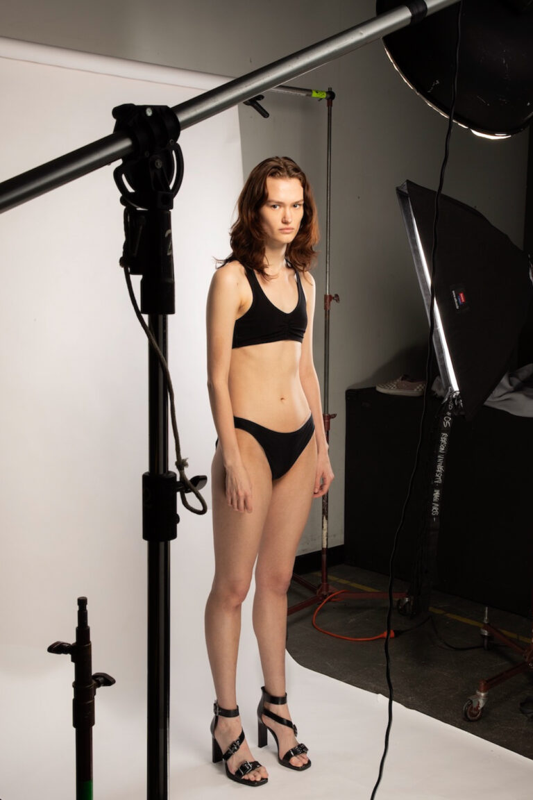 A women standing on a photo shoot set in her bra and underwear, she directly stares into the camera whilst being surrounded by various lights