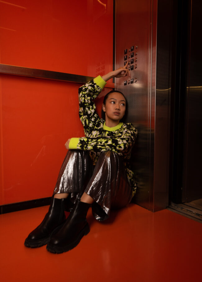 A person in shiny silver pants and a neon patterned sweater sits on the floor of a bright red elevator
