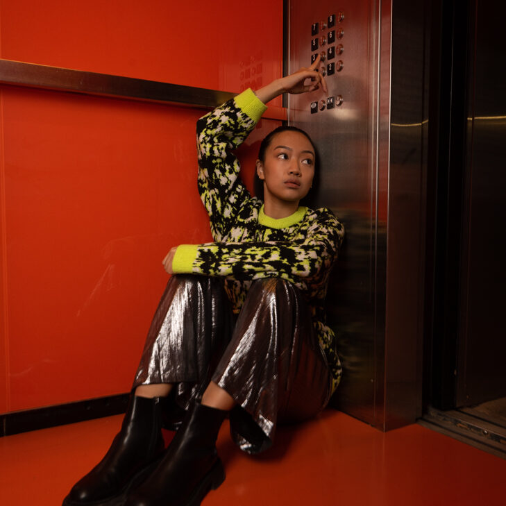 A person in shiny silver pants and a neon patterned sweater sits on the floor of a bright red elevator