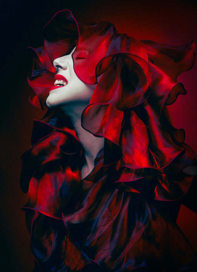 A bright red photograph of a woman expressing grief and anger. She is wearing a bright red silk garment that flows and covers her head and chest.