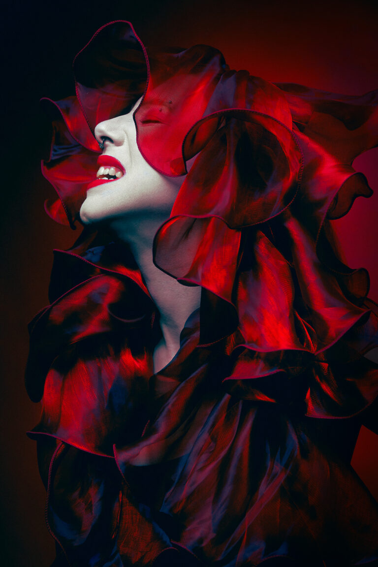 A bright red photograph of a woman expressing grief and anger. She is wearing a bright red silk garment that flows and covers her head and chest.