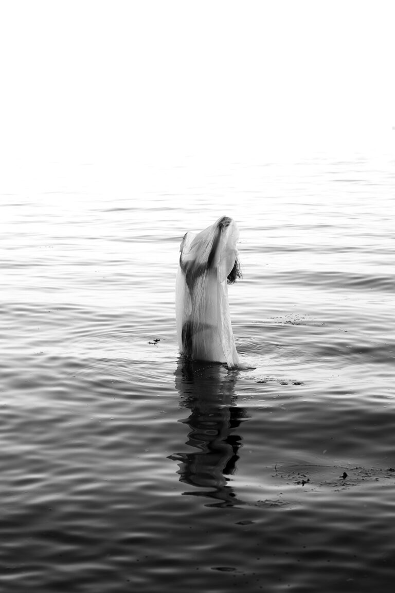 A draped figure emerges in the center of the image from a body of water. Dark tones to light tones melt into each other in a soft gradient, giving the setting a liminal feeling.