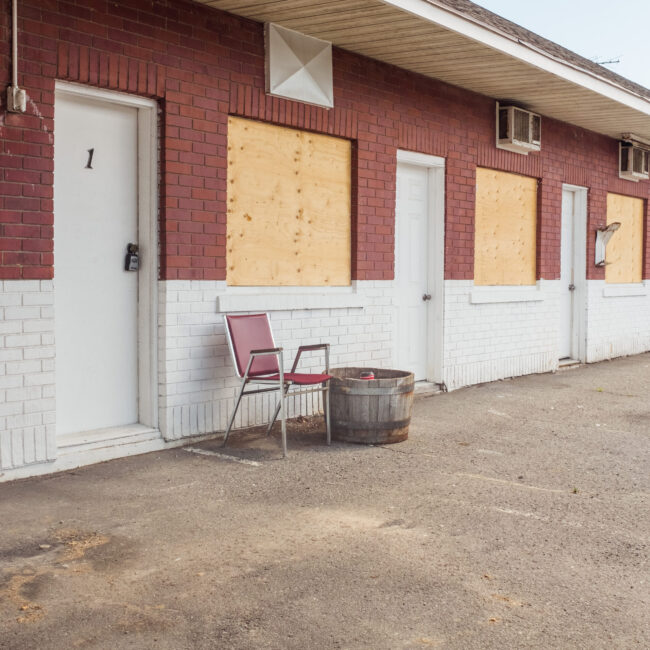 a still life image of a red chair in front of of an abandoned motel. the motel and chair has similar colour tones that makes them almost blend together.