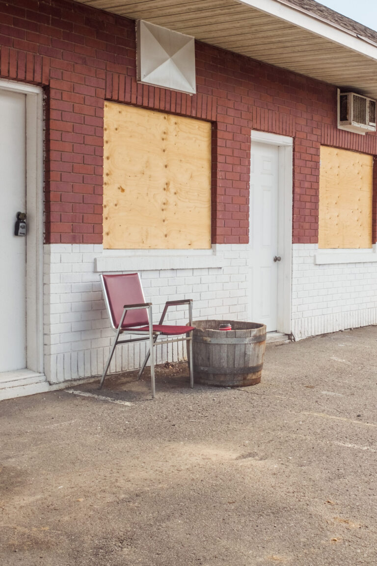 a still life image of a red chair in front of of an abandoned motel. the motel and chair has similar colour tones that makes them almost blend together.