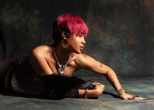 Angel: The model, Angel, is a young woman with a red short hair wearing a fur shawl and a mix of metal jewellery. In the image, she is reaching across the floor to pose and displays her accessories.