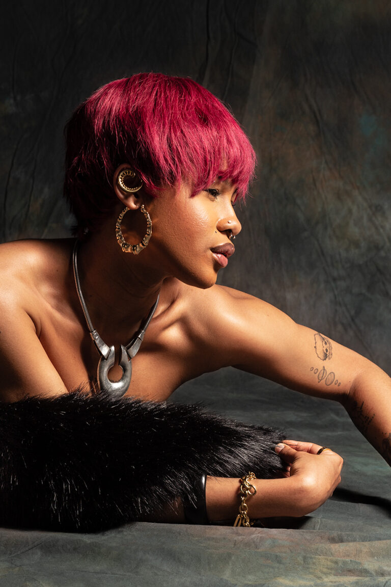 Angel: The model, Angel, is a young woman with a red short hair wearing a fur shawl and a mix of metal jewellery. In the image, she is reaching across the floor to pose and displays her accessories.